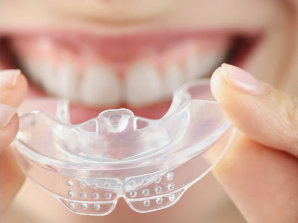 Clear dental nightguard held in front of smiling teeth, showing how a custom mouthguard protects enamel and helps reduce tooth sensitivity caused by grinding.