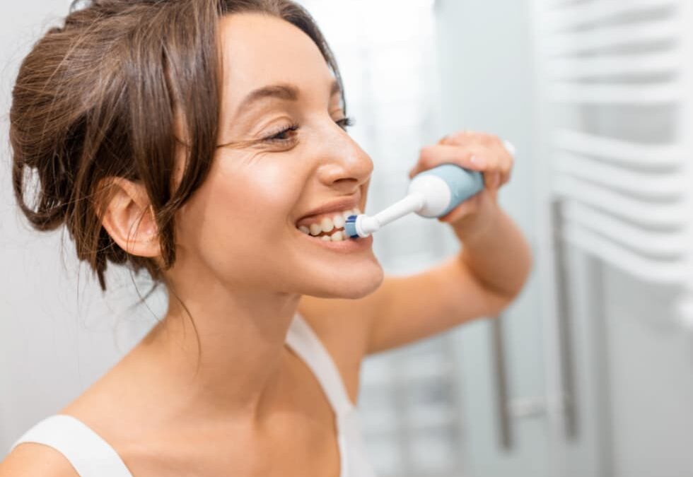 Woman smiling while brushing her teeth with an electric toothbrush, demonstrating proper daily oral hygiene.
