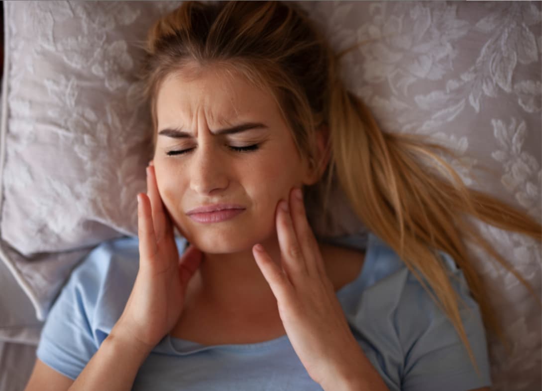 Woman waking up with jaw pain while holding both sides of her face, a common symptom of nighttime teeth grinding.