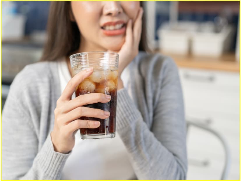 Woman wincing while holding an iced drink and touching her jaw, showing pain from tooth sensitivity.
