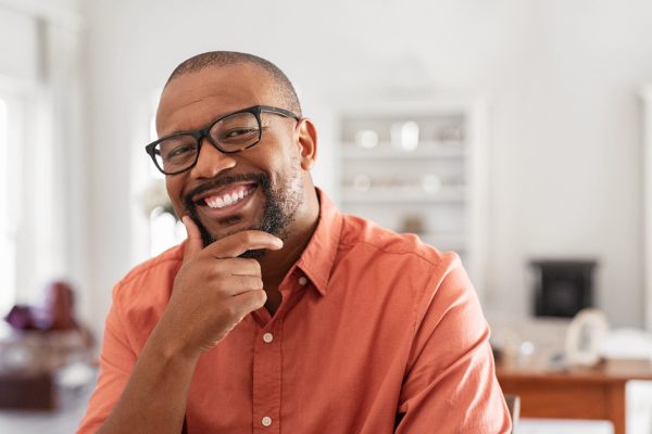 Cheerful black mature man wearing spectacles looking at camera. African businessman with hand on chin smiling while sitting at home. Happy man with eyeglasses enjoying new project.